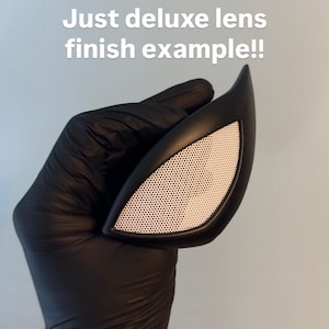 Op de afbeelding: Een zwarte, hoekige maskerlens met een wit, geperforeerd lensgedeelte. De tekst "Just deluxe lens finish example!!" wordt in het wit weergegeven. De lens wordt vastgehouden door een gehandschoende hand.