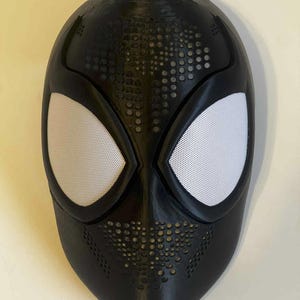 Pode incluir: Uma máscara preta com coberturas oculares de malha branca. A máscara tem uma superfície texturizada com pequenas perfurações circulares. As aberturas dos olhos são grandes e angulares, com um design distinto de aranha.