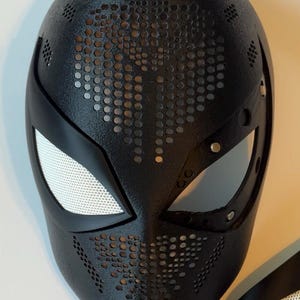 May include: Black superhero mask with a textured surface and perforated details. The mask features white mesh eye coverings and a sleek, modern design. A separate eye piece is also visible.