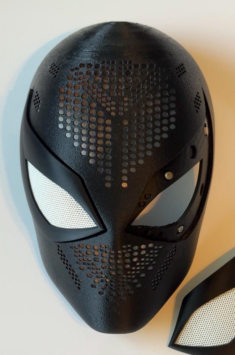 May include: Black superhero mask with a textured surface and perforated details. The mask features white mesh eye coverings and a sleek, modern design. A separate eye piece is also visible.