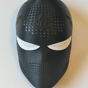 May include: A black mask with white eye coverings and a spider emblem. The mask has a textured surface with small circular perforations and is likely for cosplay or costume use.