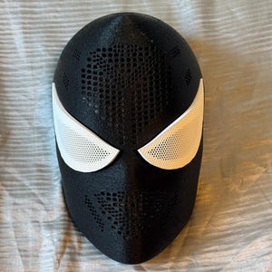 Pode incluir: Uma máscara preta com coberturas oculares brancas. A máscara tem uma superfície texturizada com pequenos orifícios. As coberturas oculares são brancas com um design perfurado. A máscara é provavelmente para cosplay ou fantasias.