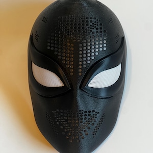 May include: A black mask with white mesh eye coverings. The mask has a textured surface with a pattern of small holes across the forehead and chin. The mask is likely for cosplay or costume use.