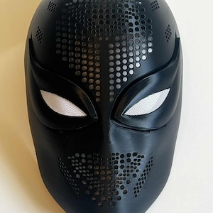 May include: A black mask with a textured surface and a design of small holes. The eye openings have a white mesh material. The mask has a pointed chin and a smooth, curved design.
