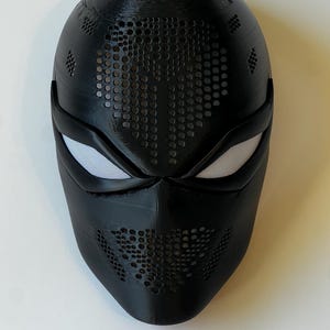 Könnte beinhalten: Eine schwarze Maske mit strukturierter Oberfläche und zahlreichen kleinen, kreisförmigen Perforationen. Die Augenöffnungen sind weiß, und die Maske hat ein schlankes, modernes Design. Die Maske ist wahrscheinlich für Kostüm- oder Cosplay-Zwecke.