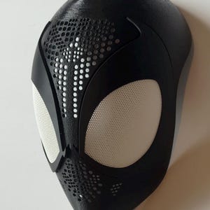 May include: A black superhero mask with white mesh eye coverings and a textured surface. The mask features a pattern of small holes and is designed to resemble a spider's web. The mask is likely for cosplay or costume use.
