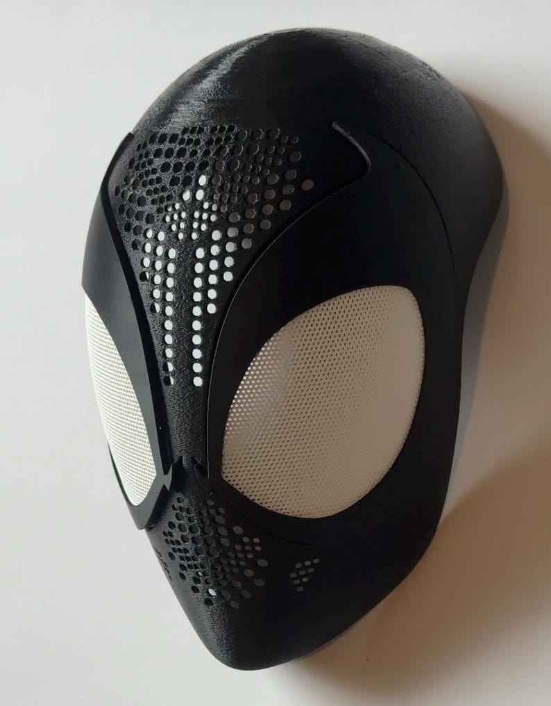 May include: A black superhero mask with white mesh eye coverings and a textured surface. The mask features a pattern of small holes and is designed to resemble a spider's web. The mask is likely for cosplay or costume use.