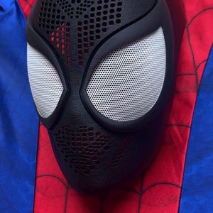 May include: A black full-face mask featuring a textured honeycomb pattern on the forehead and chin areas, with large white mesh eye lenses framed in black. The mask is positioned against a red and blue fabric background with a black web pattern.