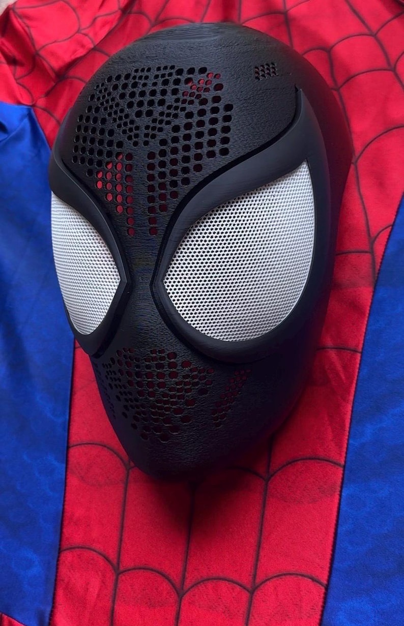 May include: A black full-face mask featuring a textured honeycomb pattern on the forehead and chin areas, with large white mesh eye lenses framed in black. The mask is positioned against a red and blue fabric background with a black web pattern.