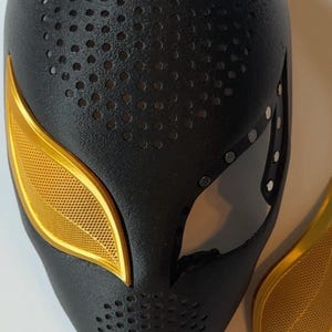 Puede incluir: M&aacute;scara de superh&eacute;roe negra y dorada con un dise&ntilde;o perforado. La m&aacute;scara presenta cubiertas y detalles dorados para los ojos. La m&aacute;scara es un accesorio de cosplay o disfraz.