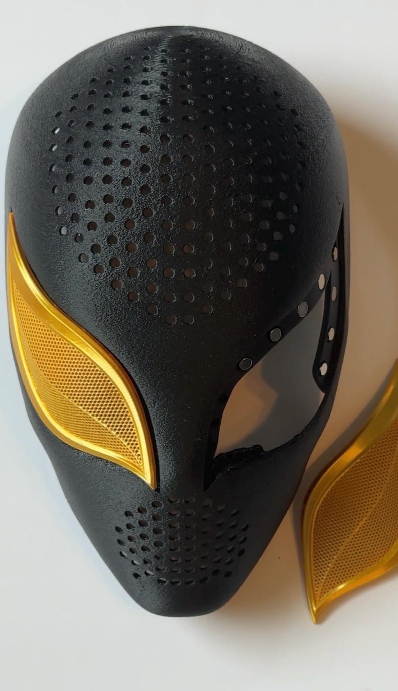 Puede incluir: M&aacute;scara de superh&eacute;roe negra y dorada con un dise&ntilde;o perforado. La m&aacute;scara presenta cubiertas y detalles dorados para los ojos. La m&aacute;scara es un accesorio de cosplay o disfraz.