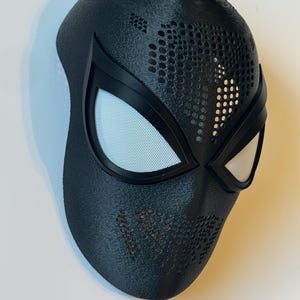 Puede incluir: Una m&aacute;scara de superh&eacute;roe negra con cubiertas oculares de malla blanca y una superficie texturizada. La m&aacute;scara tiene un dise&ntilde;o perforado en la frente y las mejillas, y probablemente est&aacute; hecha de un material duradero. La m&aacute;scara est&aacute; dise&ntilde;ada para adaptarse a la cara.