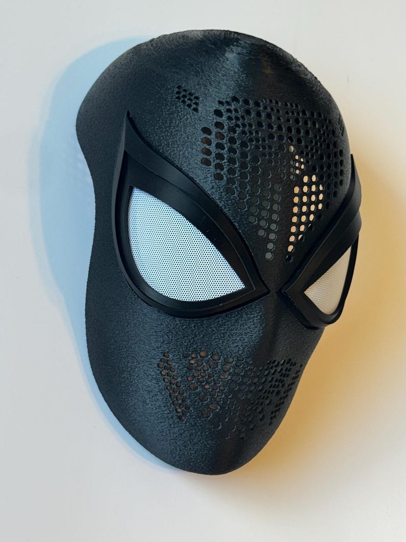 Puede incluir: Una m&aacute;scara de superh&eacute;roe negra con cubiertas oculares de malla blanca y una superficie texturizada. La m&aacute;scara tiene un dise&ntilde;o perforado en la frente y las mejillas, y probablemente est&aacute; hecha de un material duradero. La m&aacute;scara est&aacute; dise&ntilde;ada para adaptarse a la cara.