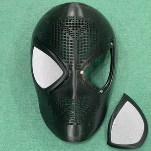 May include: A black Spider-Man mask with white mesh eye coverings and a separate matching eye piece. The mask has a textured surface with small circular perforations across the top and sides. The eye openings are outlined with a black border.
