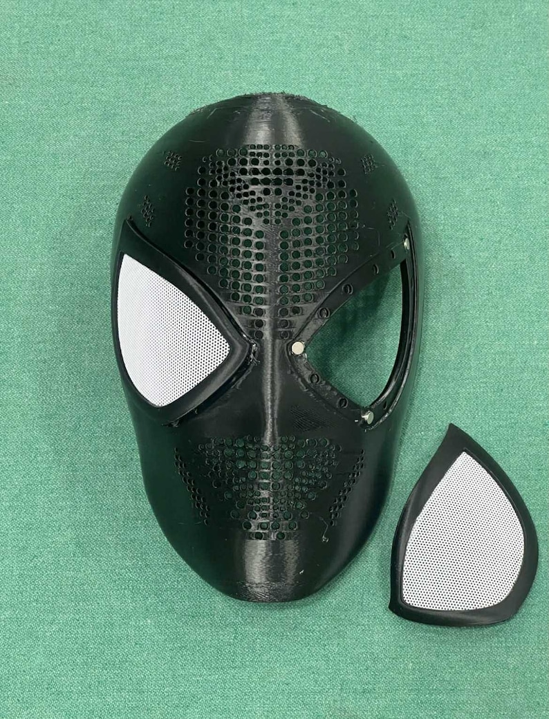May include: A black Spider-Man mask with white mesh eye coverings and a separate matching eye piece. The mask has a textured surface with small circular perforations across the top and sides. The eye openings are outlined with a black border.