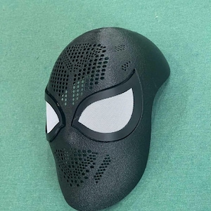 May include: Black superhero mask with white mesh eye coverings and a perforated design. The mask has a sleek, aerodynamic shape and is likely made of a durable material. The mask is designed to fit the contours of the face.