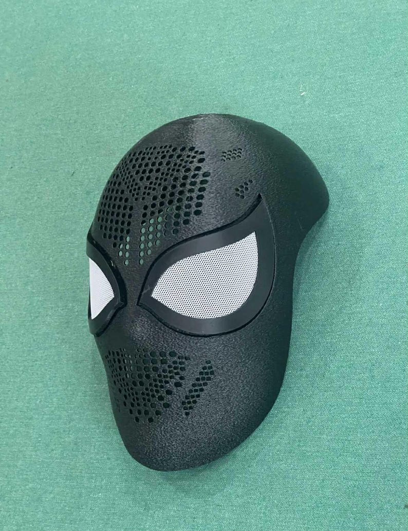 May include: Black superhero mask with white mesh eye coverings and a perforated design. The mask has a sleek, aerodynamic shape and is likely made of a durable material. The mask is designed to fit the contours of the face.