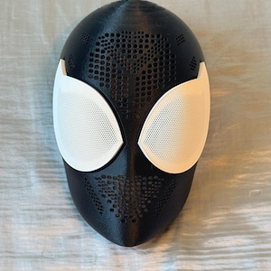 May include: A black and white Spider-Man mask with a textured surface. The mask features white eye coverings and a perforated design on the black areas. The mask is designed to cover the face.