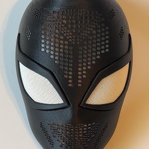 Pode incluir: Máscara preta com coberturas oculares de malha branca e superfície texturizada. A máscara tem pequenas perfurações circulares na testa e nas bochechas. Provavelmente um acessório de fantasia ou cosplay.