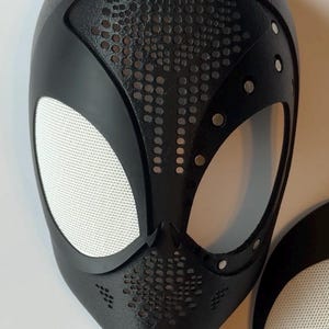 May include: Black Spider-Man mask with white mesh eye coverings. The mask has a textured surface with small circular perforations and silver rivets. A separate eye covering is also visible.