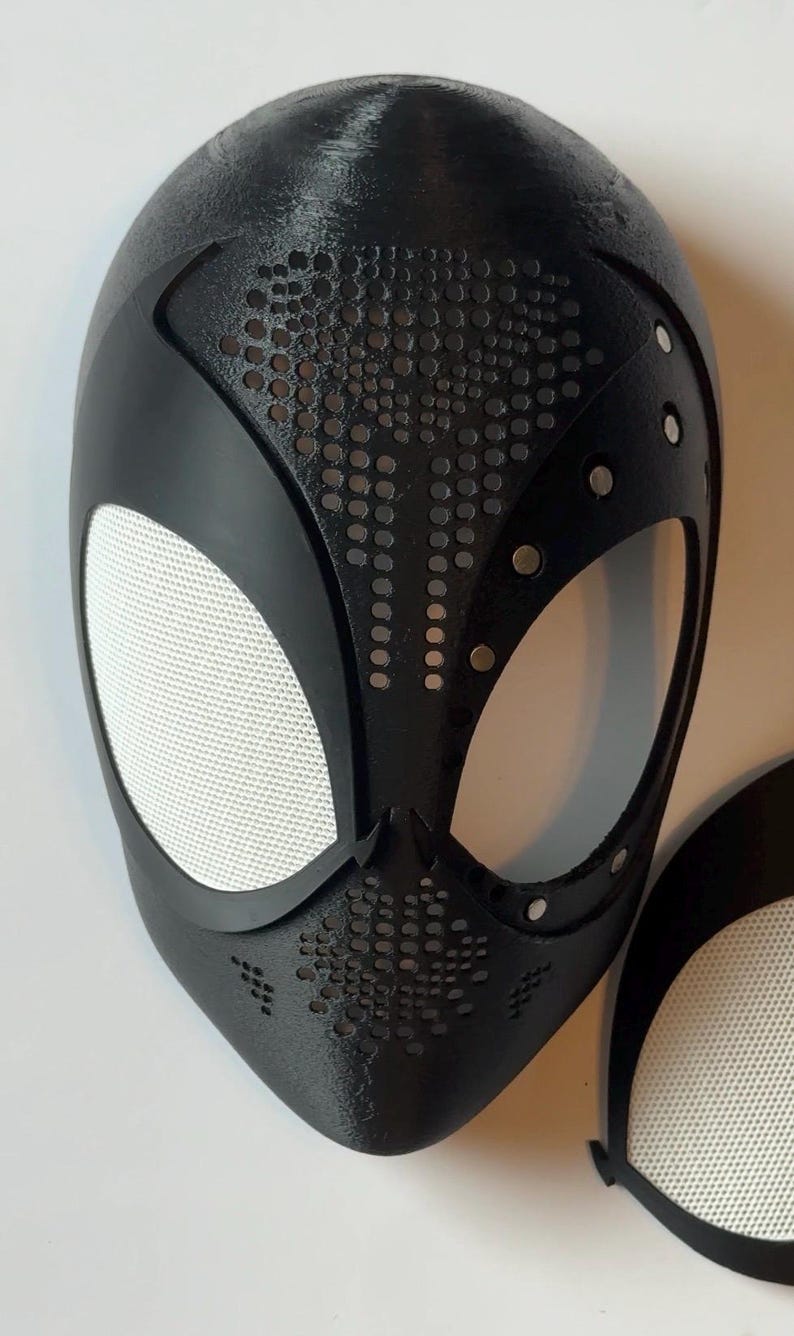May include: Black Spider-Man mask with white mesh eye coverings. The mask has a textured surface with small circular perforations and silver rivets. A separate eye covering is also visible.