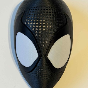 Puede incluir: Una máscara negra con cubiertas oculares de malla blanca y una superficie texturizada. La máscara tiene una serie de pequeños agujeros sobre los ojos y detalles negros alrededor de los ojos. La máscara es probablemente para cosplay o disfraces.