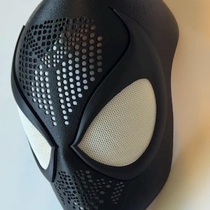 Op de afbeelding: Een zwart superheldenmasker met witte mesh oogbedekkingen en een geperforeerd ontwerp. Het masker heeft een gladde, matte afwerking en is ontworpen om de contouren van een gezicht te volgen. De oogopeningen zijn zwart omlijnd.