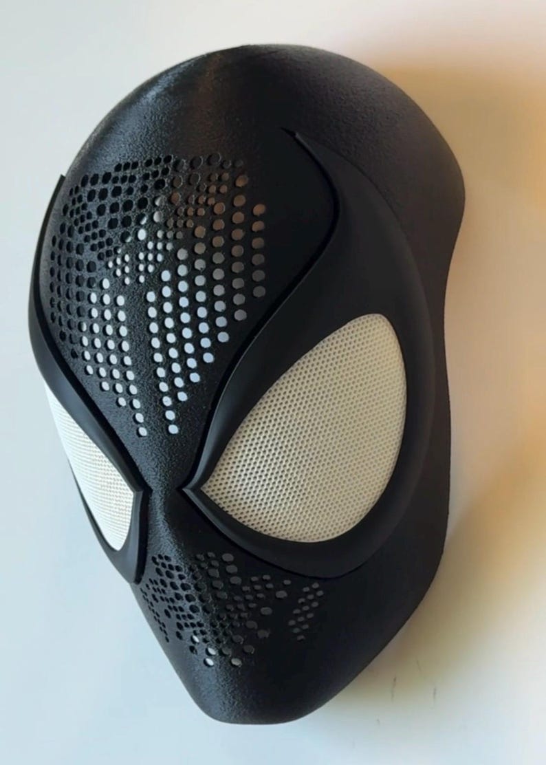 Op de afbeelding: Een zwart superheldenmasker met witte mesh oogbedekkingen en een geperforeerd ontwerp. Het masker heeft een gladde, matte afwerking en is ontworpen om de contouren van een gezicht te volgen. De oogopeningen zijn zwart omlijnd.