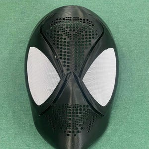 Könnte beinhalten: Eine schwarze Spider-Man-Maske mit weißen Augenabdeckungen aus Mesh. Die Maske hat eine strukturierte Oberfläche und eine Reihe kleiner Löcher um den Mundbereich. Die Maske hat eine einfarbige schwarze Farbe.