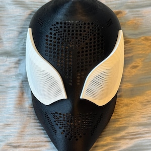 Pode incluir: Uma máscara preta e branca com uma superfície texturizada. A máscara apresenta coberturas oculares brancas e um design perfurado ao redor dos olhos e da boca. É provavelmente um acessório de fantasia ou cosplay.