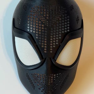May include: Black superhero mask with white mesh eye coverings and a perforated design. The mask has a textured surface and is designed to cover the entire face. The eye openings are outlined in black.