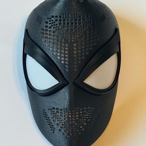 May include: A black mask with white mesh eye coverings and a textured surface. The mask has a design of small holes across the forehead and mouth area. The mask is a superhero costume accessory.