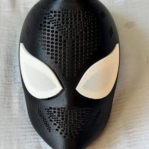 May include: A black mask with white eye coverings, featuring a perforated design across the forehead and mouth area. The mask is likely for cosplay or costume use, with a smooth, matte finish.