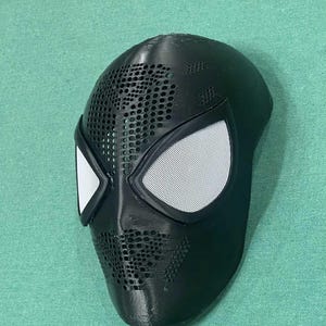 May include: Black Spider-Man mask with white mesh eye coverings and a perforated design. The mask is made of a solid material and has a smooth, curved shape. The eye coverings are outlined in black.