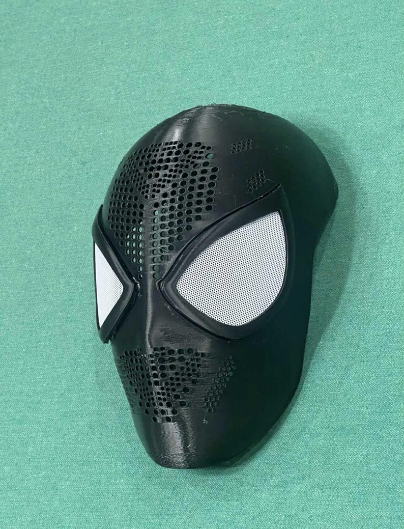 May include: Black Spider-Man mask with white mesh eye coverings and a perforated design. The mask is made of a solid material and has a smooth, curved shape. The eye coverings are outlined in black.