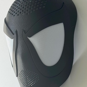 Puede incluir: Una m&aacute;scara negra con una superficie texturizada y un dise&ntilde;o estilizado. La m&aacute;scara presenta grandes aberturas para los ojos de malla blanca y peque&ntilde;os orificios de ventilaci&oacute;n. La forma general recuerda a una m&aacute;scara de superh&eacute;roe.