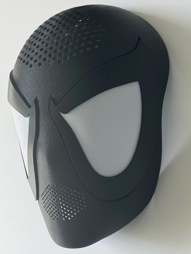 Puede incluir: Una m&aacute;scara negra con una superficie texturizada y un dise&ntilde;o estilizado. La m&aacute;scara presenta grandes aberturas para los ojos de malla blanca y peque&ntilde;os orificios de ventilaci&oacute;n. La forma general recuerda a una m&aacute;scara de superh&eacute;roe.