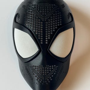 Pode incluir: Uma máscara de super-herói preta com coberturas oculares de malha branca. A máscara tem uma superfície texturizada com um padrão de pequenos orifícios e é projetada para se assemelhar ao rosto de uma aranha. A máscara é provavelmente para cosplay ou fantasia.
