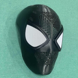May include: A black Spider-Man mask with white mesh eye coverings. The mask has a textured surface with small circular perforations across the forehead and mouth areas. The eye frames are outlined in black.