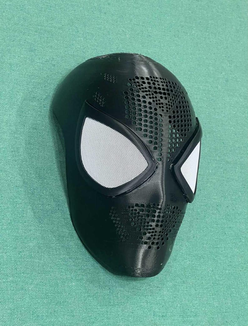 May include: A black Spider-Man mask with white mesh eye coverings. The mask has a textured surface with small circular perforations across the forehead and mouth areas. The eye frames are outlined in black.