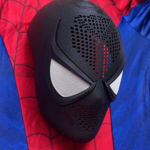 May include: A black full-face mask featuring a textured surface with perforated detailing across the forehead and mouth area. The mask includes two angular eye openings covered with white mesh lenses, framed by solid black borders.