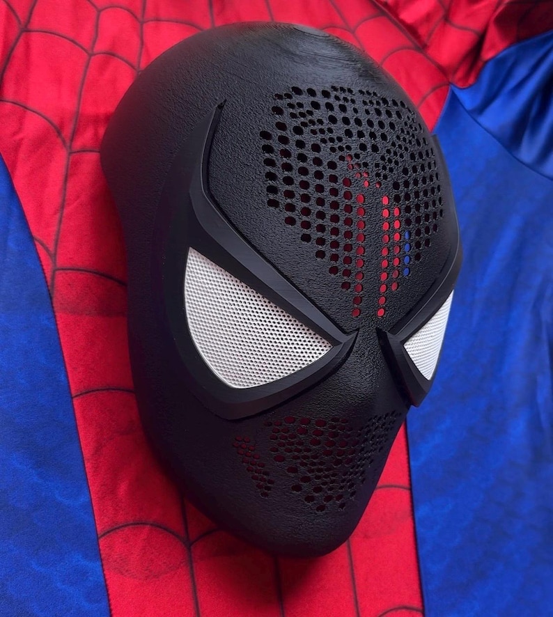 May include: A black full-face mask featuring a textured surface with perforated detailing across the forehead and mouth area. The mask includes two angular eye openings covered with white mesh lenses, framed by solid black borders.