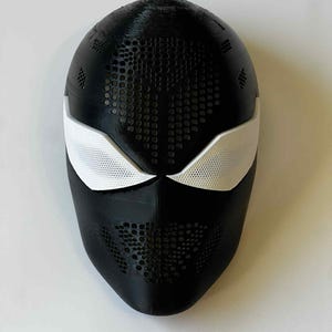 Pode incluir: Uma máscara preta e branca com uma superfície texturizada. A máscara tem coberturas brancas para os olhos e um padrão de pequenos orifícios na superfície. Provavelmente um acessório de fantasia ou cosplay.
