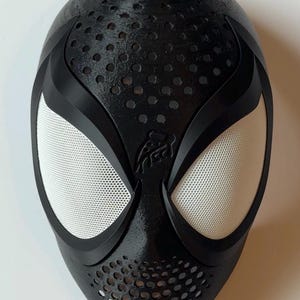 May include: Black Spider-Man mask with white mesh eye coverings and a textured surface. The mask has a series of small circular holes and a stylized spider emblem. The mask is a cosplay or costume accessory.