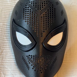 Op de afbeelding: Een zwart, volledig gezichtsmasker met een getextureerd oppervlak en een reeks kleine gaatjes voor ventilatie. De oogopeningen zijn zwart omlijnd en voorzien van witte mesh-inzetstukken. Het masker is ontworpen om op een superheldenmasker te lijken.