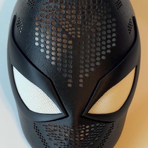 May include: Black superhero mask with white mesh eye coverings and a perforated design. The mask has a smooth, matte finish and is designed with a sleek, aerodynamic shape. The eye openings are outlined with a black border.