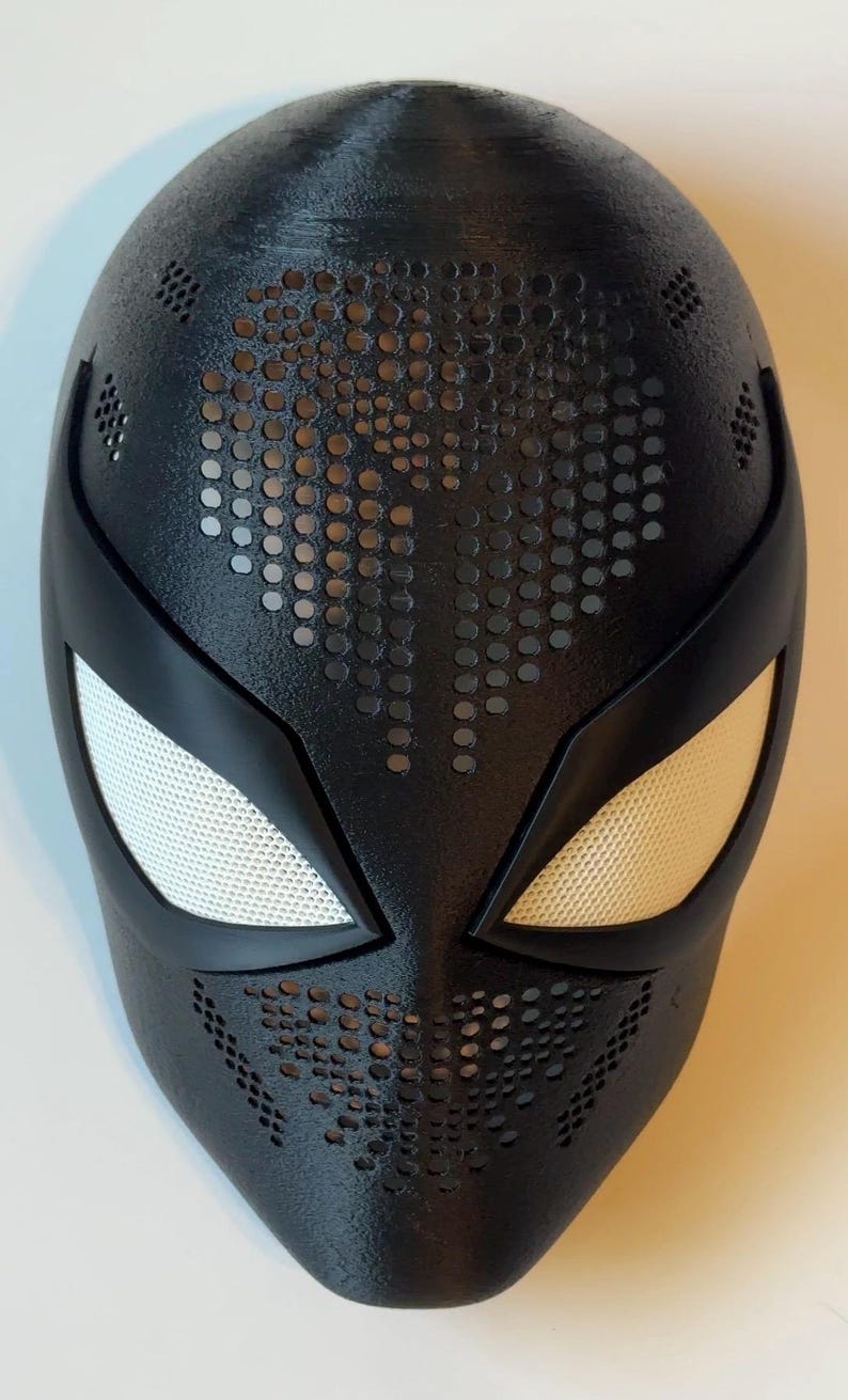 May include: Black superhero mask with white mesh eye coverings and a perforated design. The mask has a smooth, matte finish and is designed with a sleek, aerodynamic shape. The eye openings are outlined with a black border.