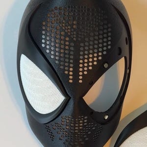 Op de afbeelding: Een zwart superheldenmasker met witte mesh oogbedekkingen en een getextureerd oppervlak. Het masker heeft een reeks kleine, gelijkmatig verdeelde gaten rond de mond en het voorhoofd. Het masker is een replica van een populair stripfiguur.