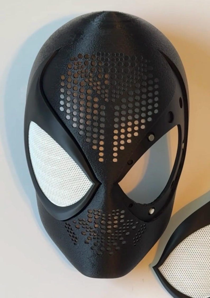 Op de afbeelding: Een zwart superheldenmasker met witte mesh oogbedekkingen en een getextureerd oppervlak. Het masker heeft een reeks kleine, gelijkmatig verdeelde gaten rond de mond en het voorhoofd. Het masker is een replica van een populair stripfiguur.