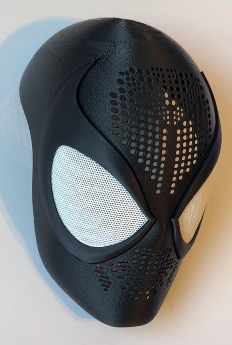 Op de afbeelding: Een zwart superheldenmasker met witte mesh oogbedekkingen. Het masker heeft een getextureerd oppervlak en is geperforeerd met kleine gaatjes rond de mond en het voorhoofd. Het is een cosplay-item.